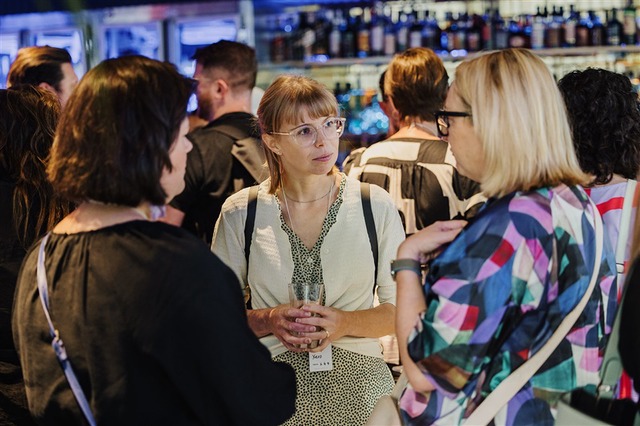 A person with glasses holds a glass and talks with others in a busy bar setting during DL23-M.