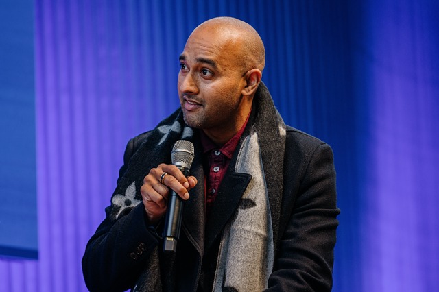Man speaking into a microphone, wearing a scarf and coat, with a blue-lit background during DO22: Reconnecting Design.